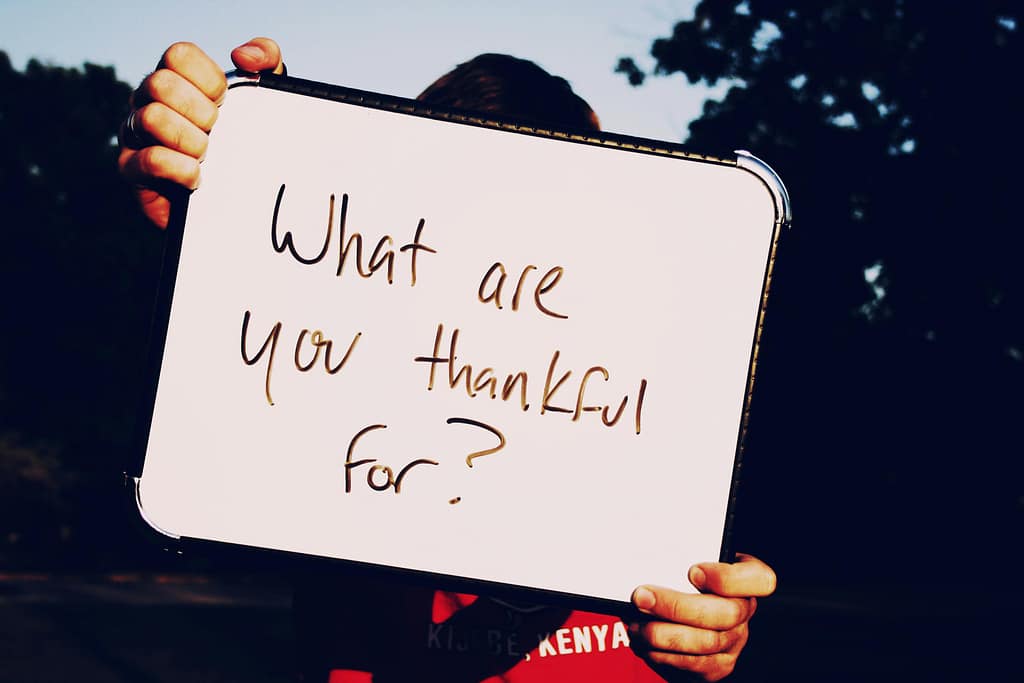 A person holds a whiteboard asking 'What are you thankful for?' against a dark outdoor background.